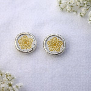 Puede incluir: Un par de pendientes de botón redondos con un diseño floral. Los pendientes presentan un fondo blanco con una flor amarilla y un borde de metal plateado. Los pendientes se muestran sobre una superficie blanca, con flores blancas en el fondo.