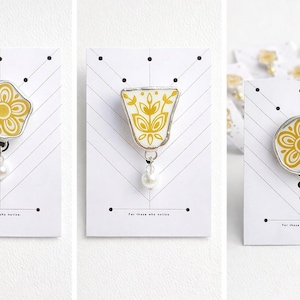 Puede incluir: Tres broches decorativos con diseños florales en amarillo y blanco, colocados sobre tarjetas blancas con un patrón de espiga. Cada broche tiene un marco plateado y una perla colgante. El texto "For those who notice." está impreso en las tarjetas.