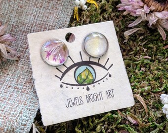 May include: A pair of silver stud earrings with a pressed flower and a white iridescent design. The earrings are displayed on a card with the text "Jewels Bright Art".