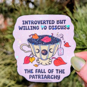 May include: A sticker with a light pink background and a teacup design. The teacup is yellow with crescent moon and star details. Text on the sticker reads "INTROVERTED BUT WILLING TO DISCUSS" and "THE FALL OF THE PATRIARCHY."