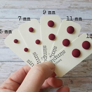 May include: Five pairs of burgundy stud earrings on white cards with the measurements 5 mm, 7 mm, 9 mm, 11 mm, and 13 mm printed on them. The cards also have the text "keep it simple" printed on them.