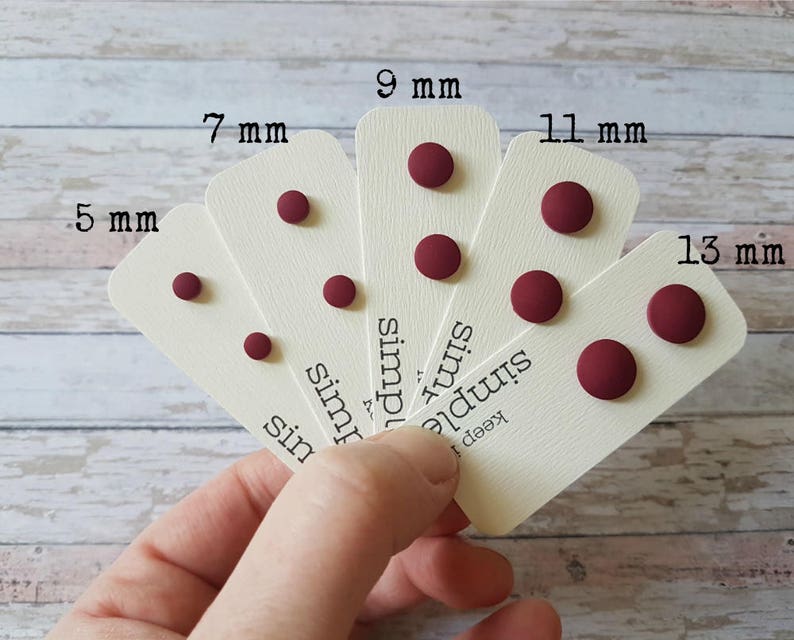 May include: Five pairs of burgundy stud earrings on white cards with the measurements 5 mm, 7 mm, 9 mm, 11 mm, and 13 mm printed on them. The cards also have the text "keep it simple" printed on them.