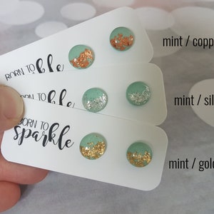 May include: A selection of mint-green stud earrings with copper, silver, and gold detailing. The earrings are circular, with metallic flakes set into the surface. They are presented on white cards, with the words "BORN TO SPARKLE" printed on them.