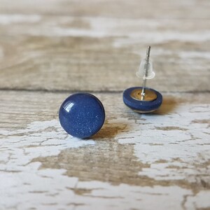 Pode incluir: Dois brincos de bot&atilde;o azul marinho. Cada brinco apresenta uma pedra redonda, brilhante, azul escuro, colocada sobre uma pequena base circular. Os pinos dos brincos s&atilde;o prateados com fechos de pl&aacute;stico transparente. Em fundo de madeira clara.