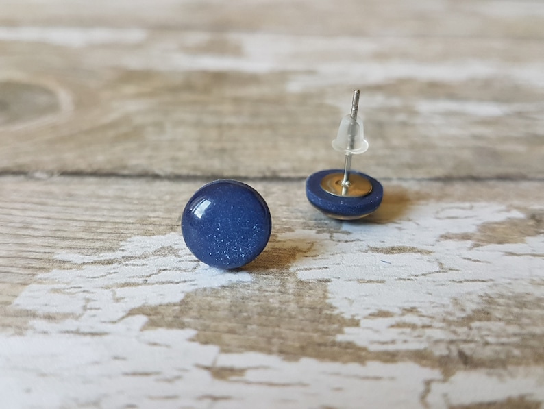 Pode incluir: Dois brincos de bot&atilde;o azul marinho. Cada brinco apresenta uma pedra redonda, brilhante, azul escuro, colocada sobre uma pequena base circular. Os pinos dos brincos s&atilde;o prateados com fechos de pl&aacute;stico transparente. Em fundo de madeira clara.