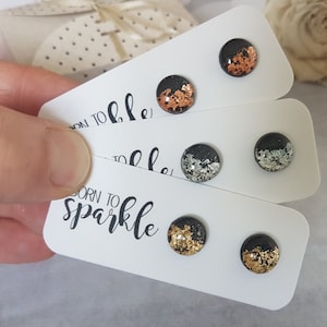 以下が含まれることがあります： 銅、銀、金色のメタリック箔アクセントが付いた黒いスタッドピアス3組のクローズアップ。各イヤリングは、黒い筆記体で「BORN TO sparkle」というテキストが書かれた白いカードに表示されています。