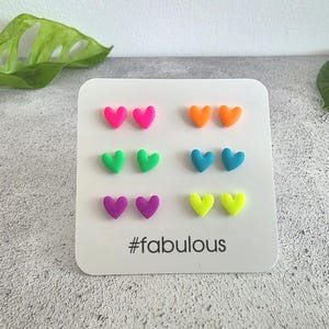 May include: A set of six pairs of small heart-shaped stud earrings in neon pink, orange, bright green, teal, purple, and yellow, displayed on a white square card with rounded corners featuring the text #fabulous printed in black at the bottom.