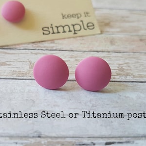 Op de afbeelding: Twee ronde roze oorbel studs met stalen of titanium pinnen. De oorbellen liggen op een houten oppervlak met een kaartje waarop "keep it simple" staat.