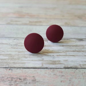 May include: Two round, burgundy-colored stud earrings on a light wood surface.