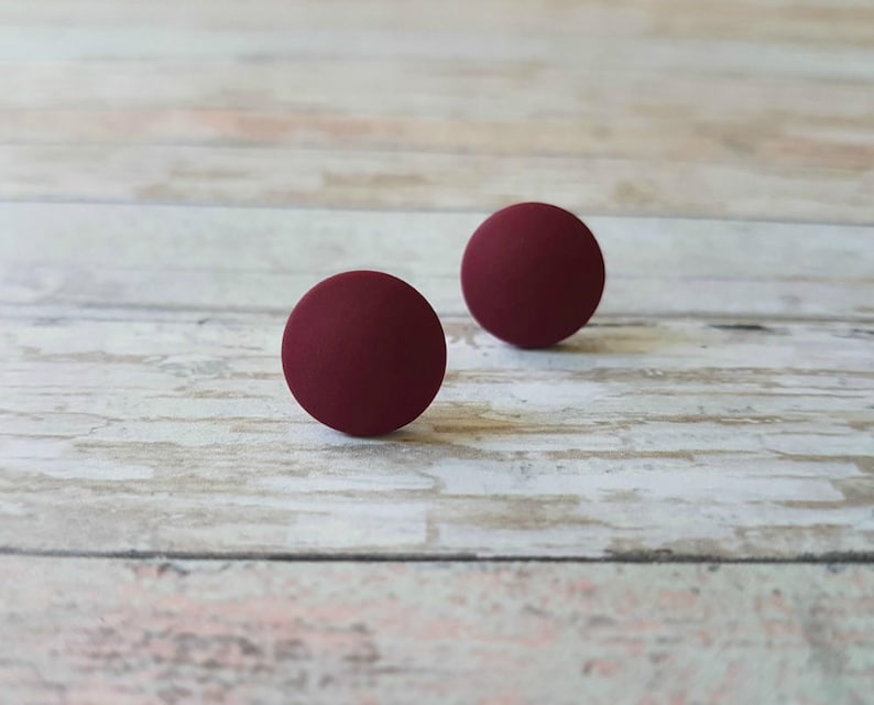 May include: Two round, burgundy-colored stud earrings on a light wood surface.