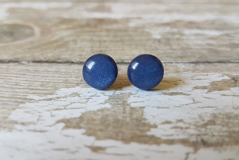 Pode incluir: Dois brincos redondos azuis escuros com uma apar&ecirc;ncia salpicada. Os brincos s&atilde;o colocados sobre um fundo claro de gr&atilde;o de madeira, criando um contraste que real&ccedil;a a sua cor e forma. Um acess&oacute;rio simples, mas elegante.