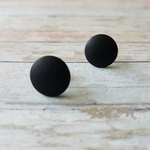 May include: Two black round stud earrings on a white wooden surface.