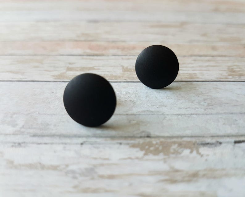 May include: Two black round stud earrings on a white wooden surface.