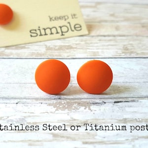 Può includere: Un paio di orecchini rotondi arancioni con perni in acciaio inossidabile o titanio. Gli orecchini sono esposti su una superficie di legno bianco con un biglietto che dice "keep it simple".