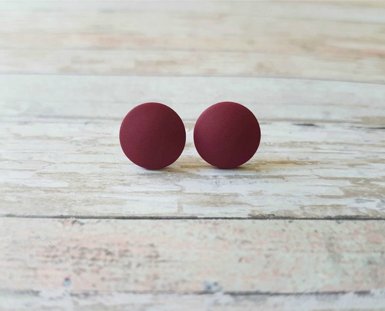 May include: A pair of round, burgundy stud earrings. The earrings are made of a smooth, matte material.