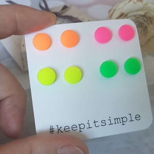Op de afbeelding: Een set van zes neonkleurige oorstekers in oranje, roze, groen en geel, gerangschikt op een witte kaart. De kaart heeft de tekst "#keepitsimple" in zwart gedrukt.