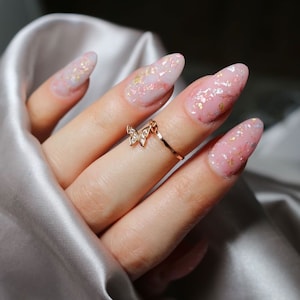 Pode incluir: Close-up de uma mão usando um anel de ouro com um amuleto de borboleta. As unhas estão pintadas com um esmalte de unha rosa claro com detalhes de glitter dourado.