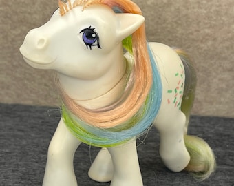 May include: A white pony toy with a rainbow mane and tail. The pony has a purple eye and a pink and green confetti pattern on its side.