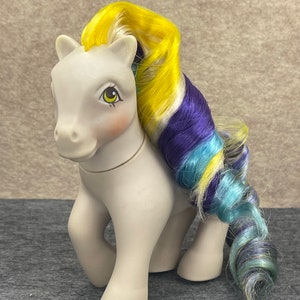 Könnte beinhalten: Ein graues My Little Pony mit gelbem, blauem und violettem Haar. Das Pony hat braune Augen und einen rosa Rouge auf den Wangen.