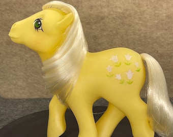 May include: A yellow toy pony with a white mane and tail. The pony has green eyes and white floral designs on its side. The toy is standing on a dark surface.