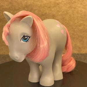 May include: A gray toy pony with pink hair and a pink tail. The pony has blue eyes and pink heart-shaped markings on its side. The toy is a vintage collectible.