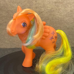 Puede incluir: Un poni de juguete naranja con una melena y cola fluidas en tonos de amarillo, verde y azul. El poni tiene marcas moradas en forma de estrella en su costado y pequeñas alas. El poni está sobre una superficie negra.