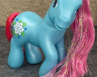 Può includere: Un giocattolo di pony blu chiaro con criniera e coda rosa e bianca. Il pony ha un fiore bianco dipinto sul fianco.