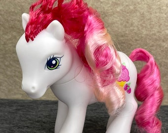 May include: White pony with pink and white curly hair. The pony has a blue eye with green accents and a pink and yellow cupcake with strawberries on its flank.