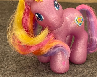 May include: A pink glittery My Little Pony toy with a yellow, pink, and purple mane and tail. The pony has a blue heart with a sunburst design on its flank.