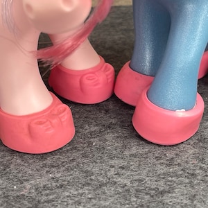 May include: Close-up of toy pony legs. The legs are light pink and blue, with pink hooves. The hooves have a detailed design. The background is a grey surface.