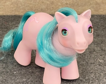 May include: A pink My Little Pony toy with blue hair and large eyes. The pony has a purple ribbon on its flank.