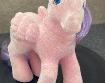 Könnte beinhalten: Ein Vintage-Pony-Spielzeug aus rosa Plüsch mit blauen Augen und lila Haaren. Das Pony hat kleine Flügel und steht auf einer schwarzen Oberfläche. Das Spielzeug ist ein klassisches Sammlerstück.