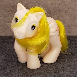 Puede incluir: Un pony blanco con pelo amarillo y un ala rosa con estrellas. El pony tiene ojos azules con pupilas amarillas.
