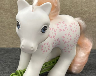 Könnte beinhalten: Ein weißes My Little Pony Spielzeug mit einer rosa Mähne und einem rosa Schwanz. Das Pony hat rosa und weiße geometrische Muster auf seinem Körper und eine grüne Feder, die an seinen Beinen befestigt ist.
