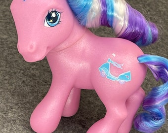 Può includere: Un pony giocattolo rosa con capelli blu e viola. Il pony ha una grafica di scooter blu sul lato e dettagli a forma di stella negli occhi. I capelli del pony sono acconciati a riccioli.