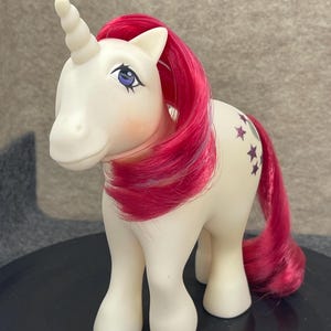 May include: A cream-colored unicorn toy with a spiraled horn, bright pink mane and tail, and purple eyes. The unicorn features a graphic design on its flank consisting of several small, dark purple stars arranged in a curved pattern.