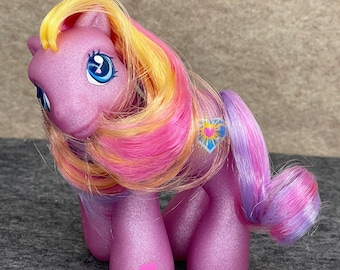 Può includere: Un pony viola glitterato con un cuore rosa sulla sua gamba. Il pony ha una lunga criniera multicolore con striature gialle, rosa e viola.