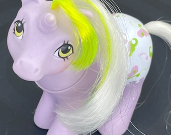 May include: A purple unicorn My Little Pony toy with a yellow and white mane and tail. The pony is wearing a white and green floral print dress.