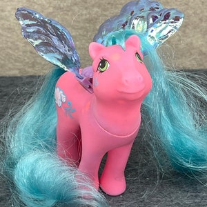May include: Pink My Little Pony with blue hair and iridescent wings. The pony has a floral design on its side and is standing on a gray surface.