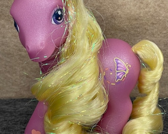 May include: A purple My Little Pony with long, yellow, iridescent hair. The pony has a butterfly on its flank and a pink heart on its hindquarters.