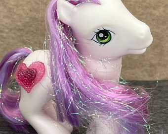 May include: A white pony with a purple and pink iridescent mane and tail. The pony has a pink heart-shaped symbol on its flank.