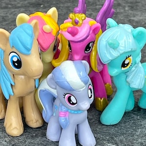 May include: Five small plastic pony figurines. The ponies are in various colors, including pink, blue, and purple. They have different colored manes and tails, and some have horns. The ponies are all standing on a gray surface.