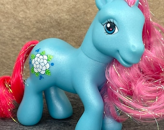 May include: A light blue pony toy with pink hair and a white flower design on its side. The pony has blue eyes and a pink nose.