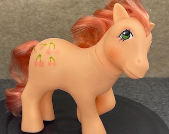 Op de afbeelding: Een vintage perzik-kleurig speelgoedpony met een roze manen en staart. De pony heeft groene ogen en een kersendesign op zijn flank. Het speelgoed staat op een zwart oppervlak.