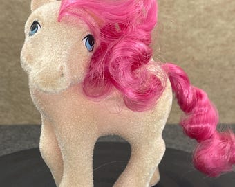 May include: A vintage toy pony with a light tan body and bright pink hair. The pony has blue eyes and is standing on a black surface. The hair is styled in a curly fashion, and the tail is also pink and curly.