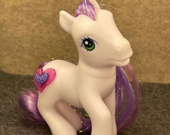 May include: A white pony with a purple and pink heart on its flank. The pony has a purple mane and tail with glitter accents.