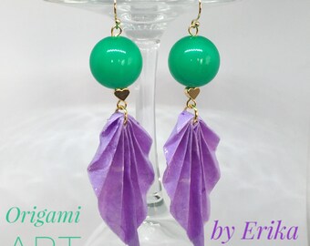 Può includere: Orecchini in origami viola con perline rotonde verdi e dettagli dorati. Gli orecchini sono appesi a un supporto in vetro. Il testo "Origami ART by Erika TRIOSCHI" è visibile nell'immagine.