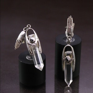 May include: Two silver-toned earrings. Each earring features a clear crystal pendant, a Yin Yang symbol, and a small hand charm. The earrings are displayed on black cylindrical stands against a dark background.