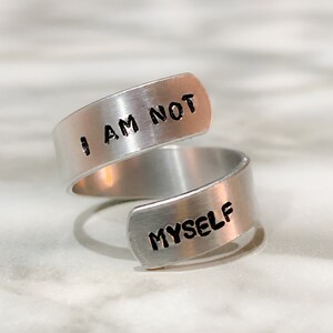 wrap style silver coloured ring with the words "i am not" on the top band and "myself" on the bottom band. Words are coloured black.