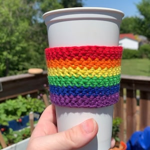 Puede incluir: Un vaso blanco para llevar con un calentador de taza de ganchillo arcoíris. El calentador está hecho con hilo rojo, naranja, amarillo, verde, azul y morado.